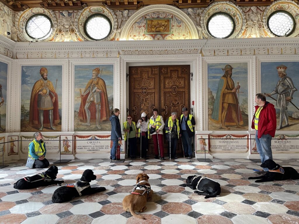 5 Führhunde sind im Spanischen Saal abgelegt, Die Menschen sind vor einer alten geschnitzten grossen Holztüre für das Foto aufgestellt. Der Boden ist aus Marmorplatten, es sind Gemälde von Rittern auf die Wandverkleidung gemalt.