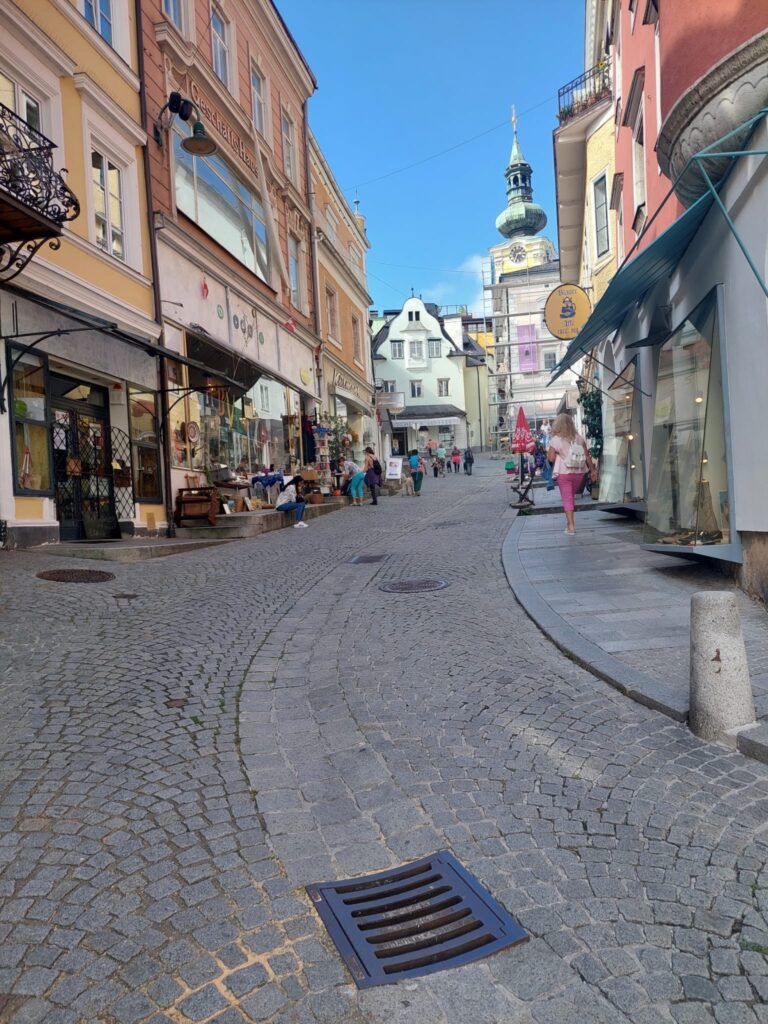 An uphill cobblestone street leading into the old city of Gmunden. The guide dog teams are almost invisible but there is a blue sky and an old church tower with an onion roof.